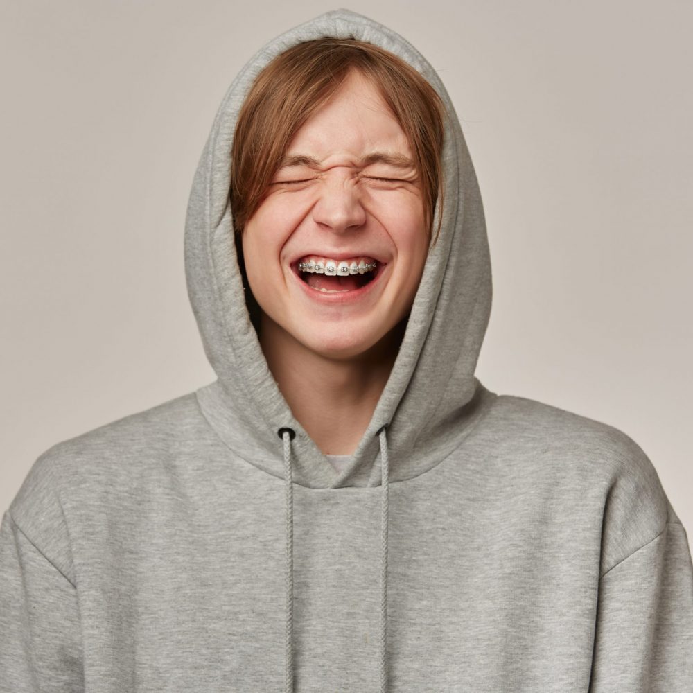 Cheerful male, handsome guy with blond hair. Wearing grey hoodie. Has braces. People and emotion concept. Puts hood on and laughing with closed eyes. Stand isolated over grey background